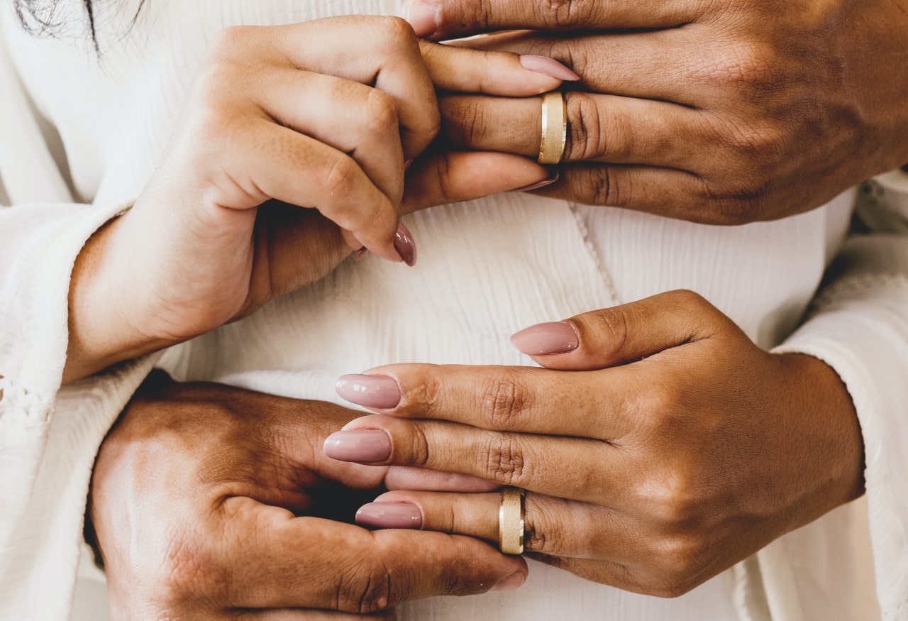 A man and woman simultaneously sliding matching wedding bands onto each other’s fingers.