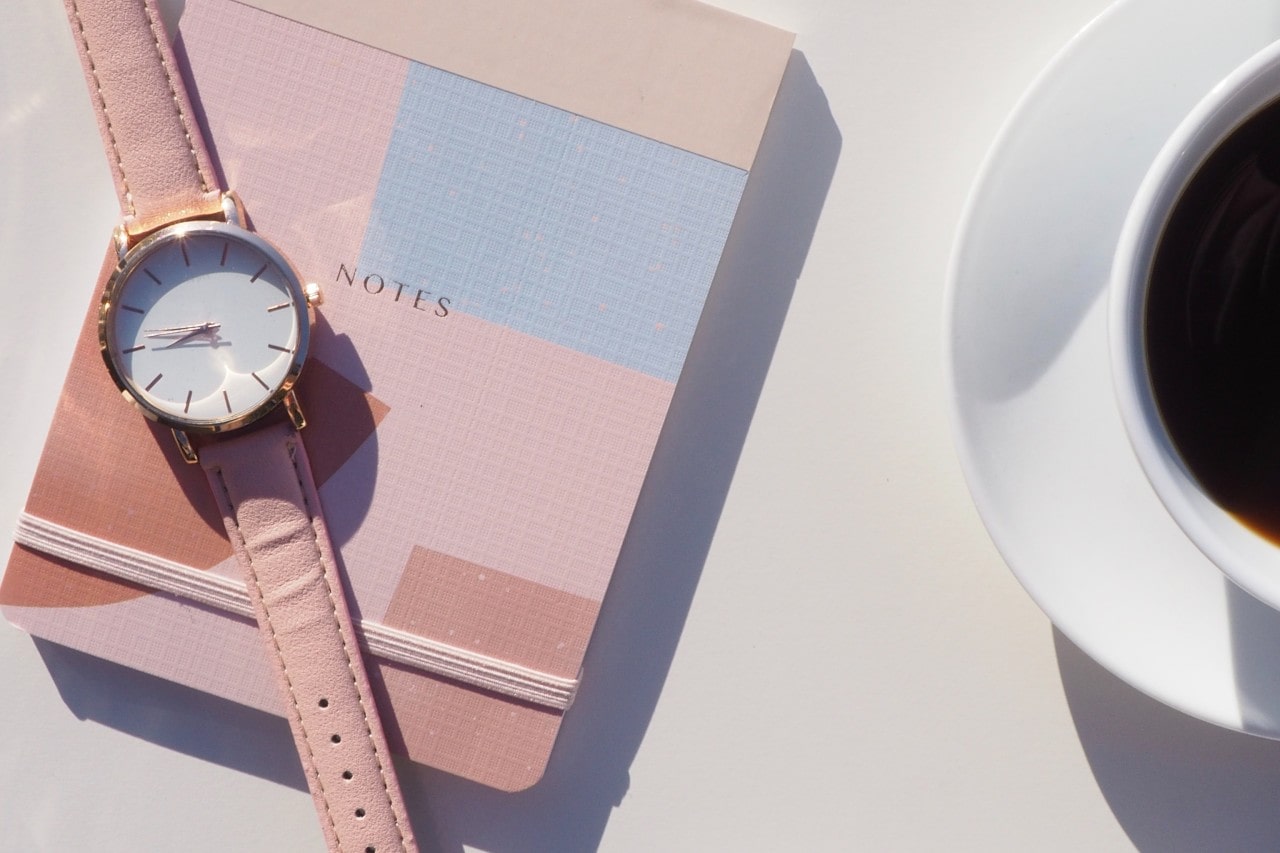 A minimalist watch with a pink leather band displayed atop a notebook, next to a cup of coffee.