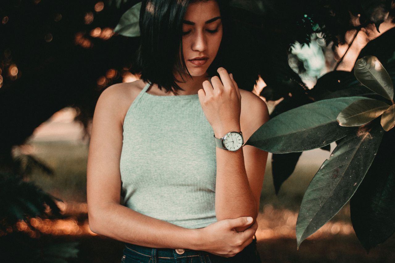 A casually dressed woman wearing a fashionable watch in a leafy area.