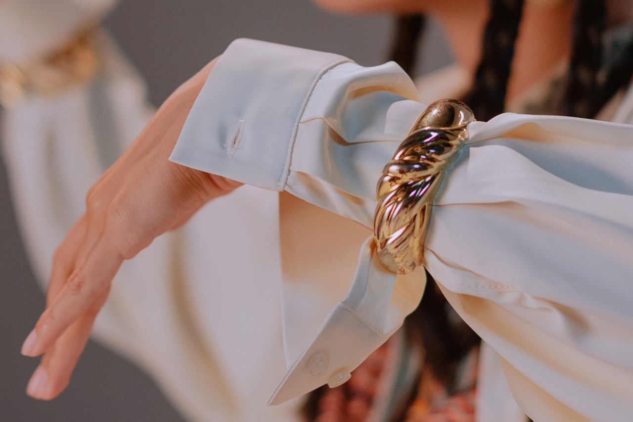 a woman’s arm adorned with a white button up sleeve and a bold yellow gold bangle bracelet