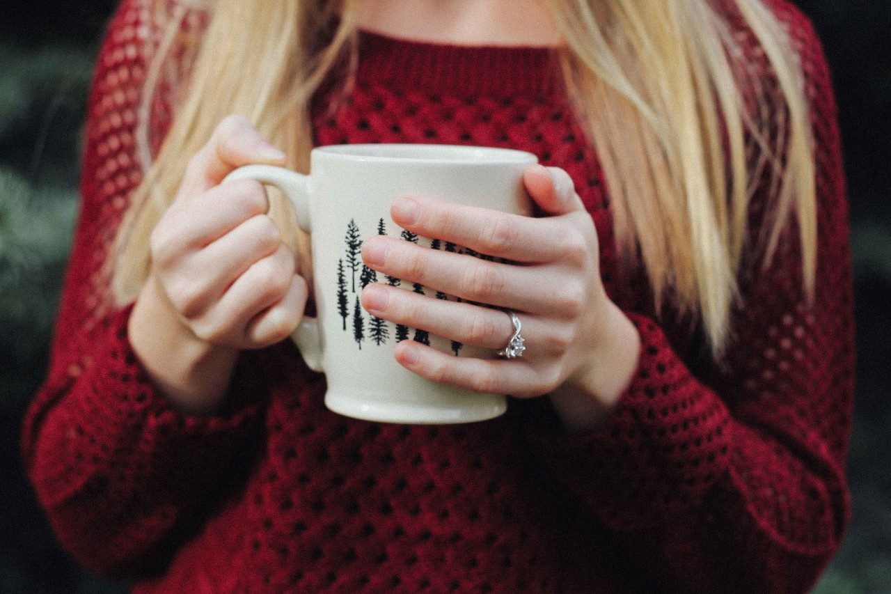A woman in a sweater sips coffee out of a mug, wearing a twisted shank ring.
