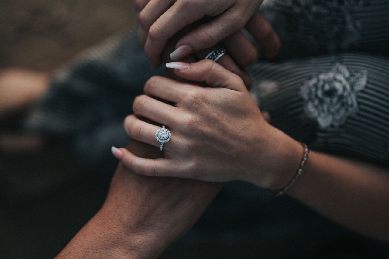 A woman holds a man’s hands wearing a halo engagement ring.