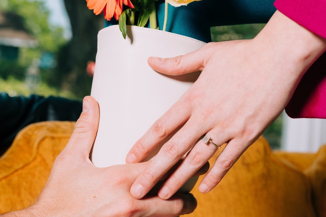 a couple’s hands passing a flower pot, the woman wearing a solitaire engagement ring