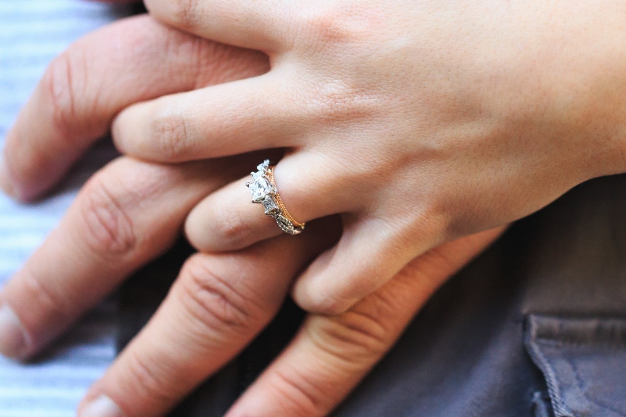 a woman’s hand over a man’s–the woman’s hand adorned with a mixed metal three stone ring