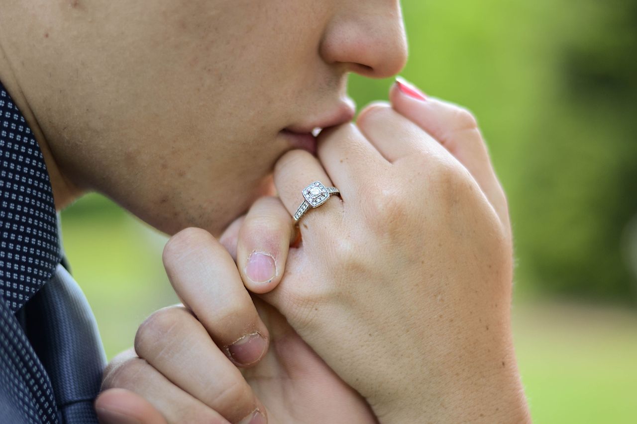 a man lifting his fiance’s hand to his lips–her finger adorned with a halo engagement ring