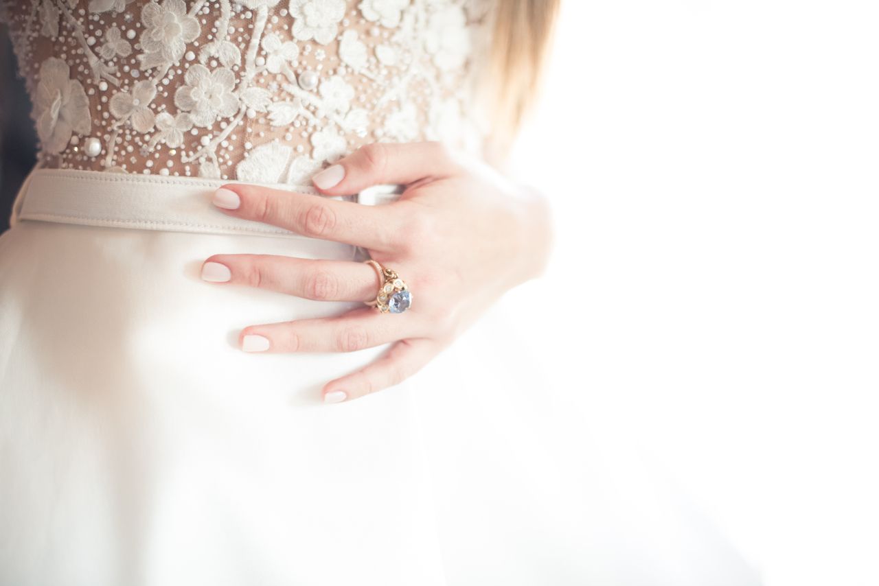 A bride’s hand with a gold ring featuring a vibrant gemstone, resting on her lace wedding gown