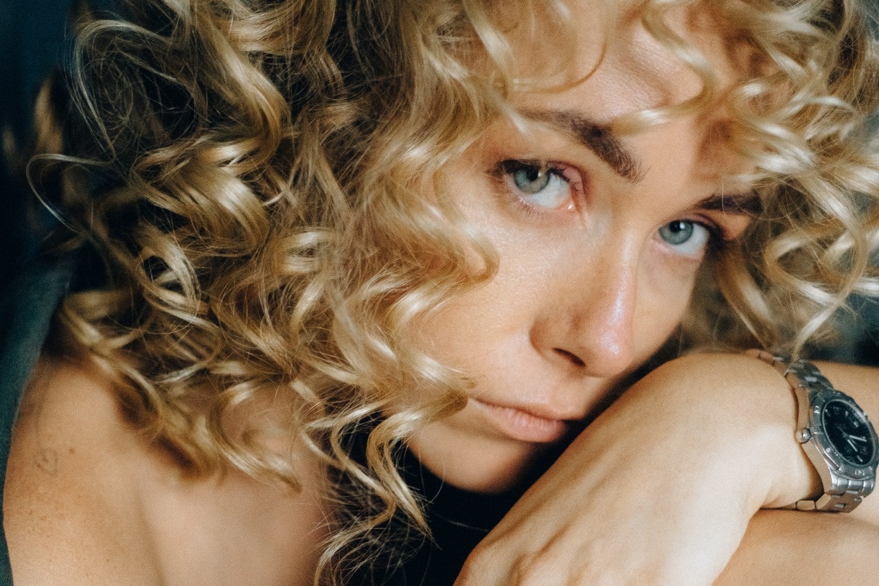 A close-up of a young woman with curly hair wearing a luxury watch.